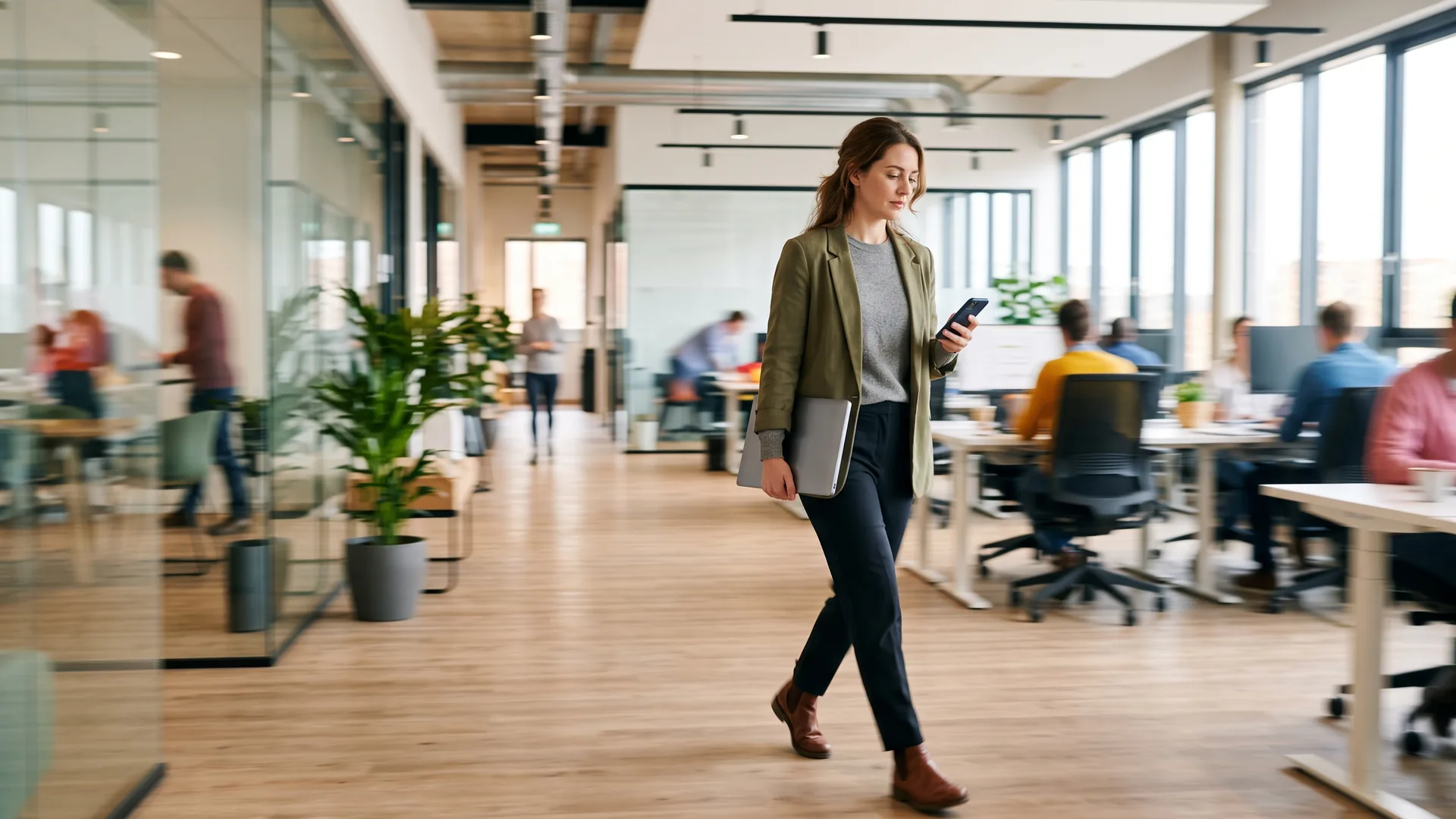 A woman strides through a busy sunlit open-plan office checking her phone, a laptop under her arm, as colleagues work in soft blur behind her.